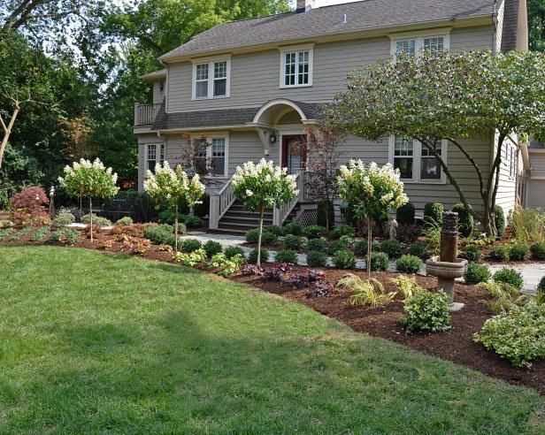 A beige, two-story house with a manicured front garden featuring four small flowering trees and a stone fountain.