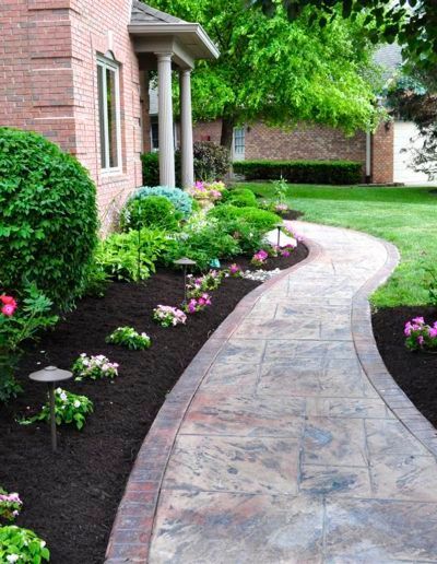 A curved stamped concrete walkway leads to the brick entryway of a home, bordered by garden beds with shrubs and flowers.