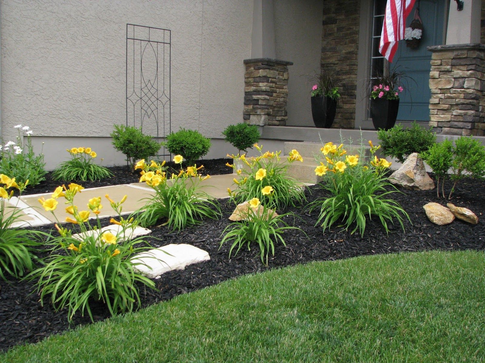 A landscaped front garden bed with yellow daylilies, green shrubs, and stone pavers leading to a house entrance.