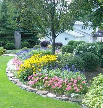 A lush garden bed with pink and yellow flowers, green shrubs, and a tree, bordered by stones against a white house.