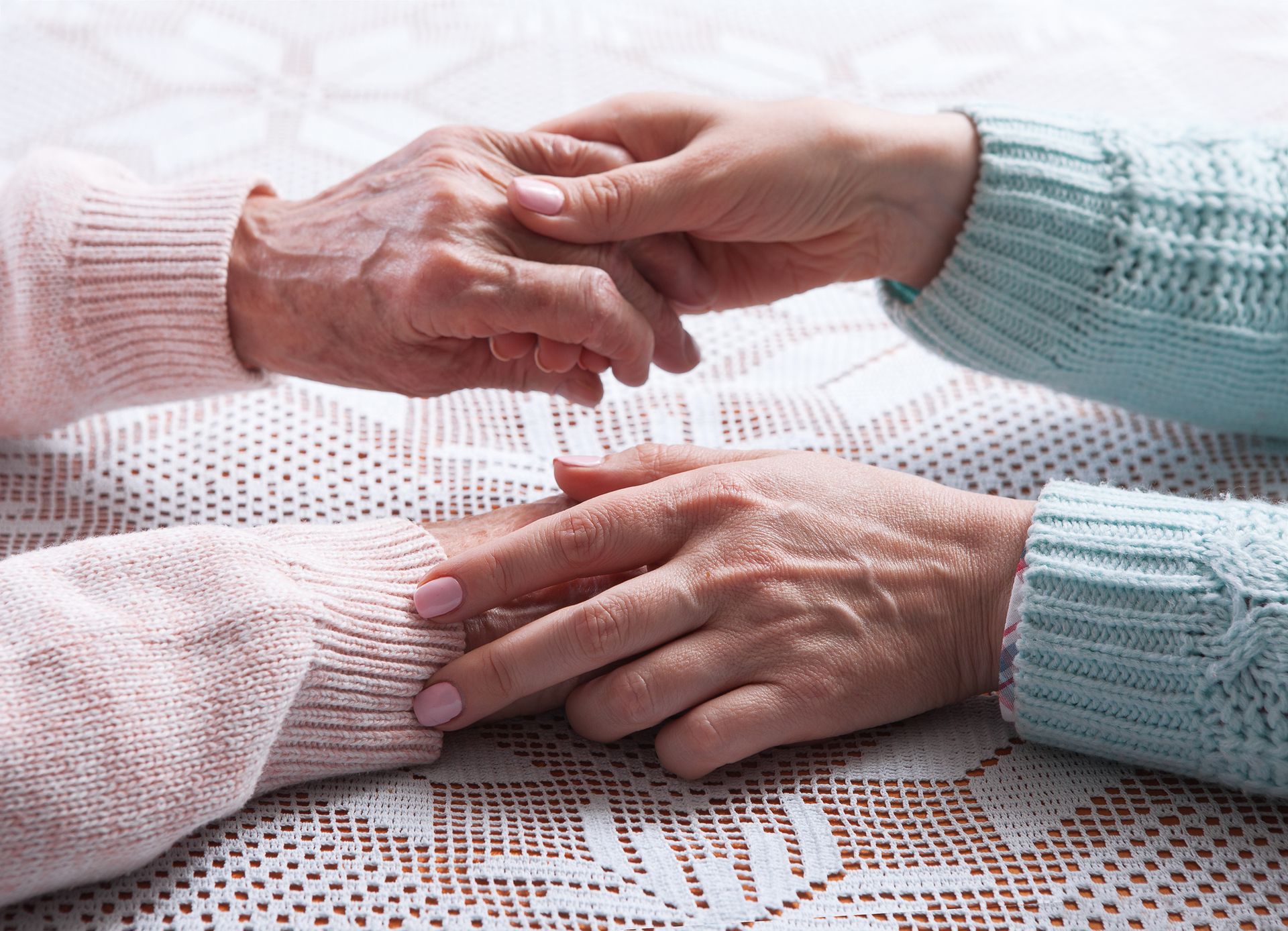 Elderly and young hands clasped in support over lace tablecloth, symbolizing care and connection.