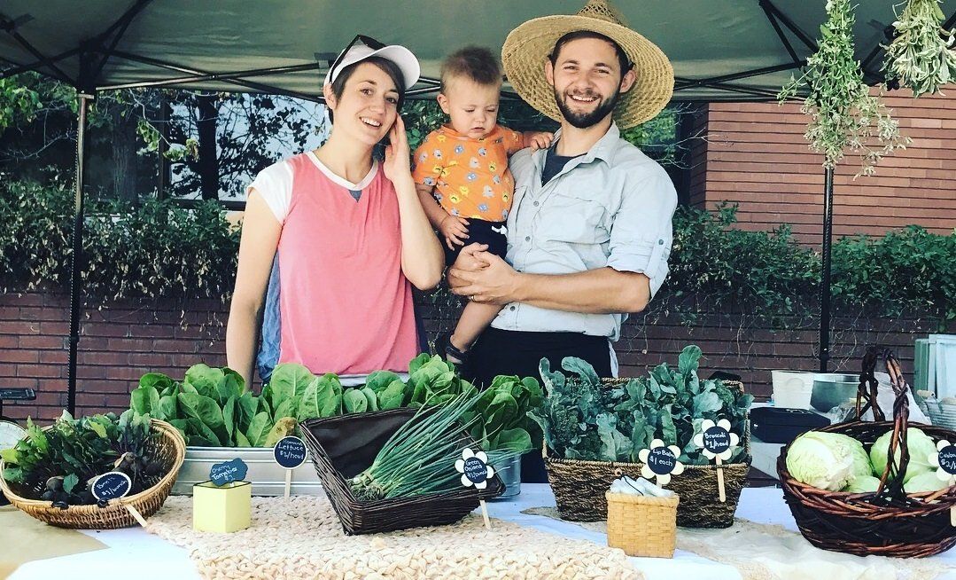 A man and woman are holding a baby in front of a table with vegetables.