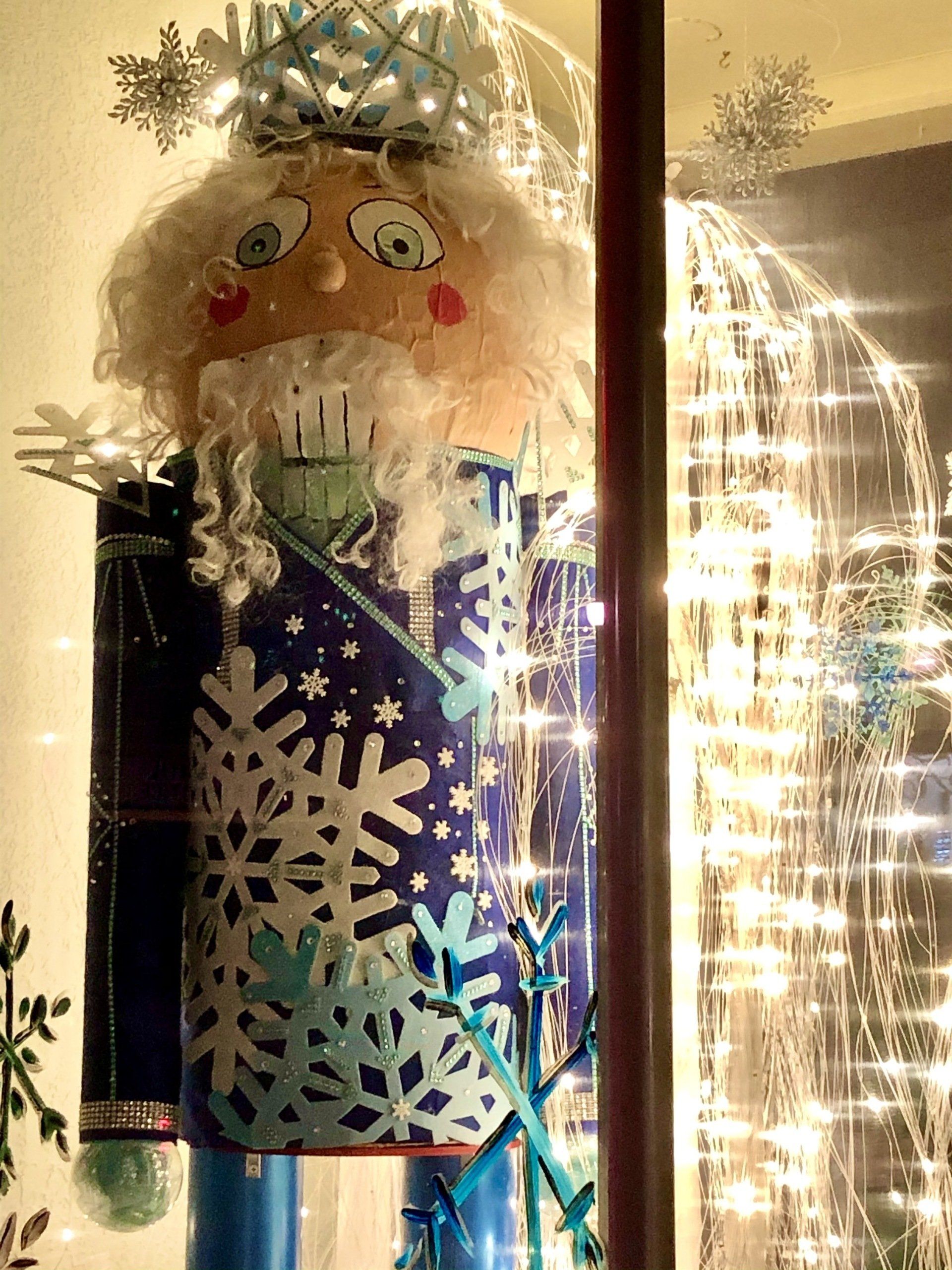 A nutcracker is standing in front of a window with christmas lights behind him.