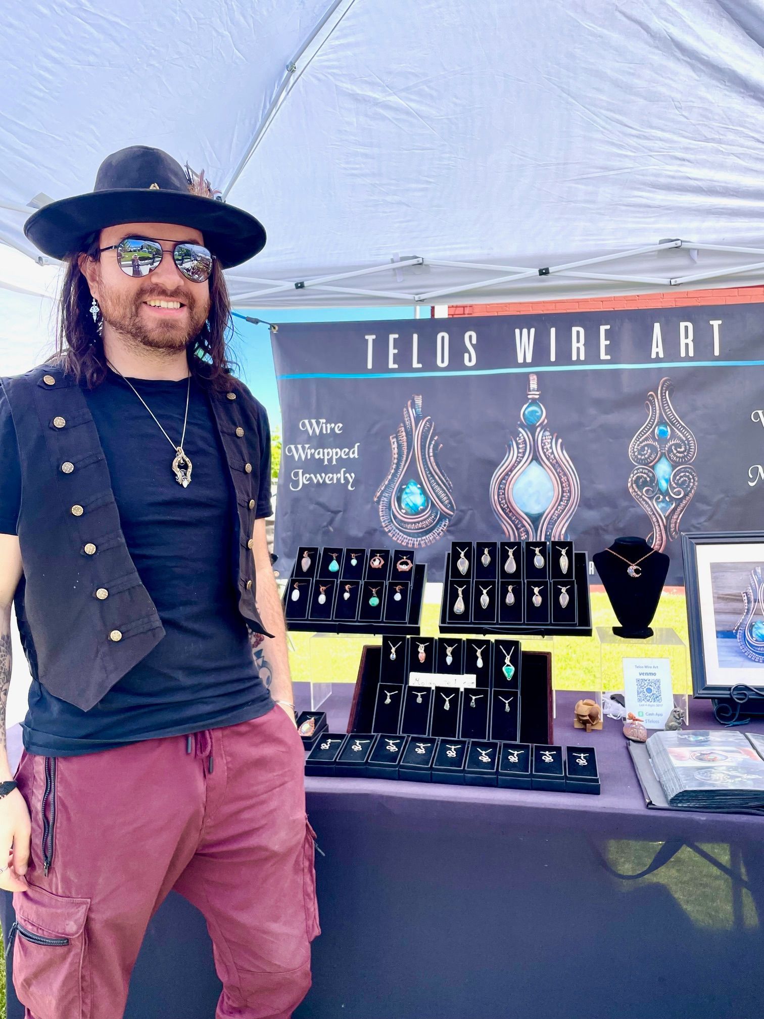 A man wearing a hat and sunglasses is standing in front of a table with jewelry on it.