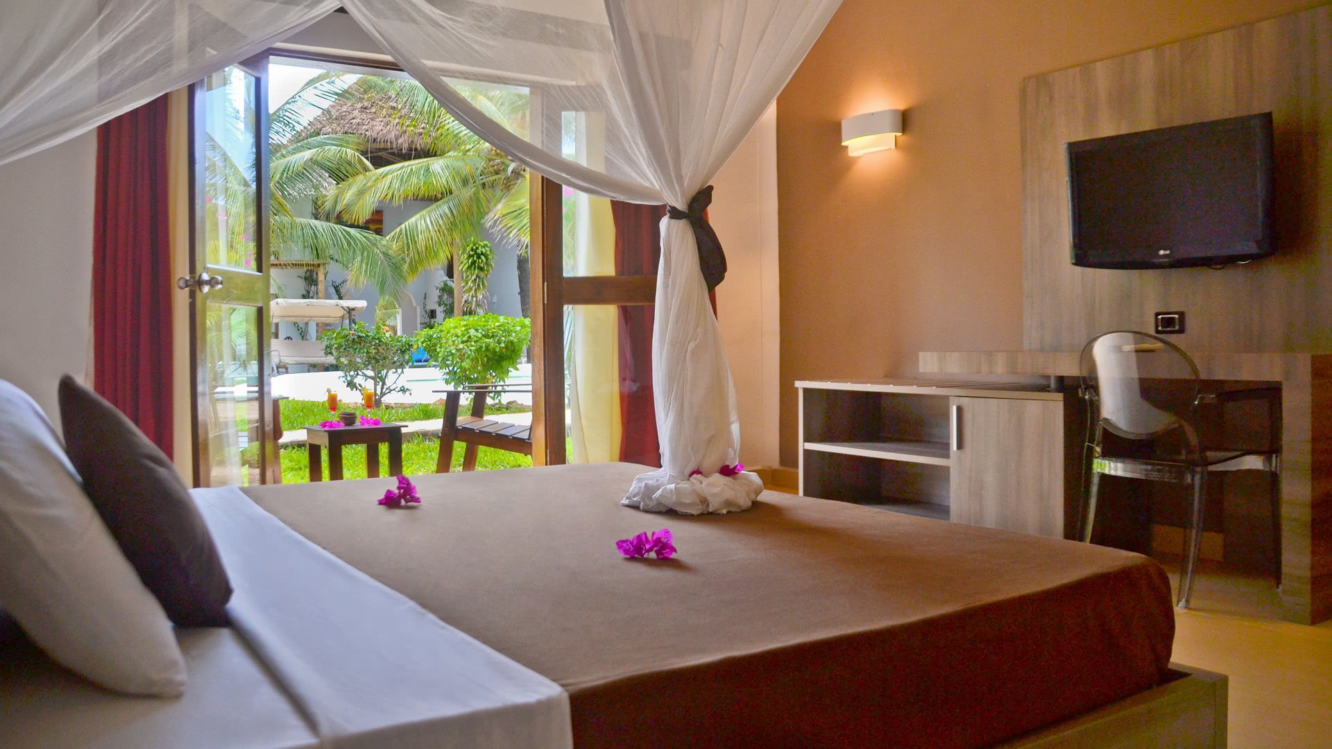 Camera da letto con letto, porta aperta sul giardino, TV, scrivania. Colori tenui marrone e bianco.