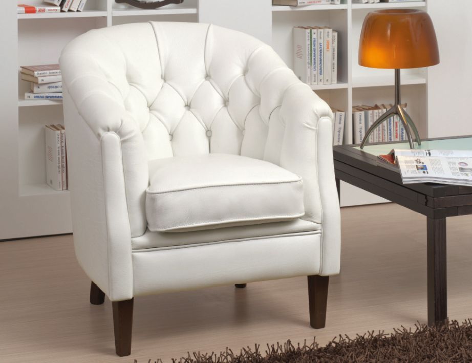 Poltrona bianca imbottita con gambe in legno scuro, cuscino bianco, lampada e libri sullo sfondo.