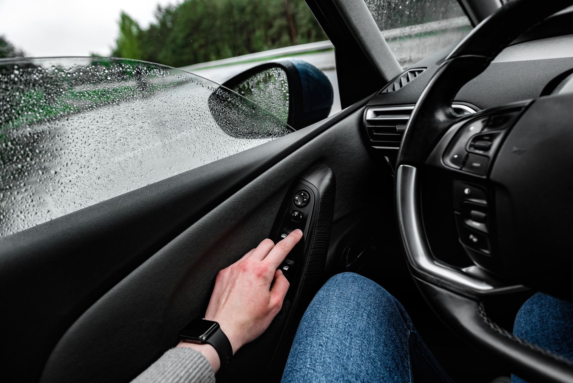 Person inside a car, adjusting the side mirror control on a rainy day.