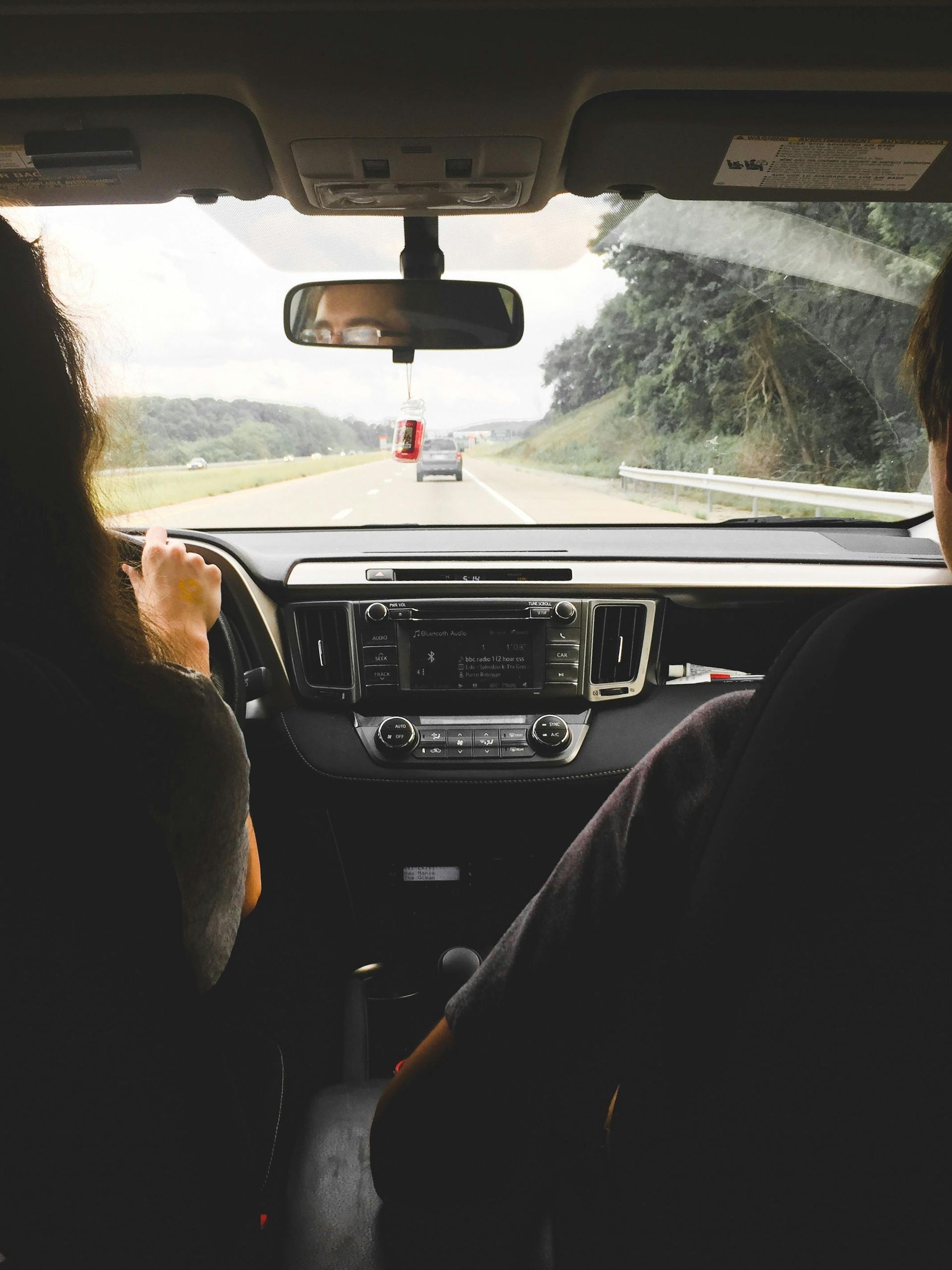 Inside a car, person driving on a highway. Passenger visible. Trees on the right, other cars in the distance.