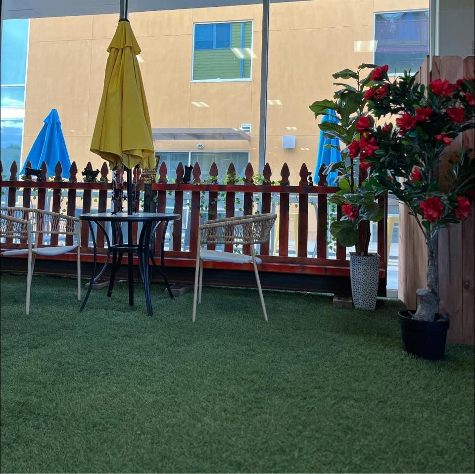 Outdoor cafe seating area with umbrella, table, chairs, and potted red flowering tree against a wooden fence.