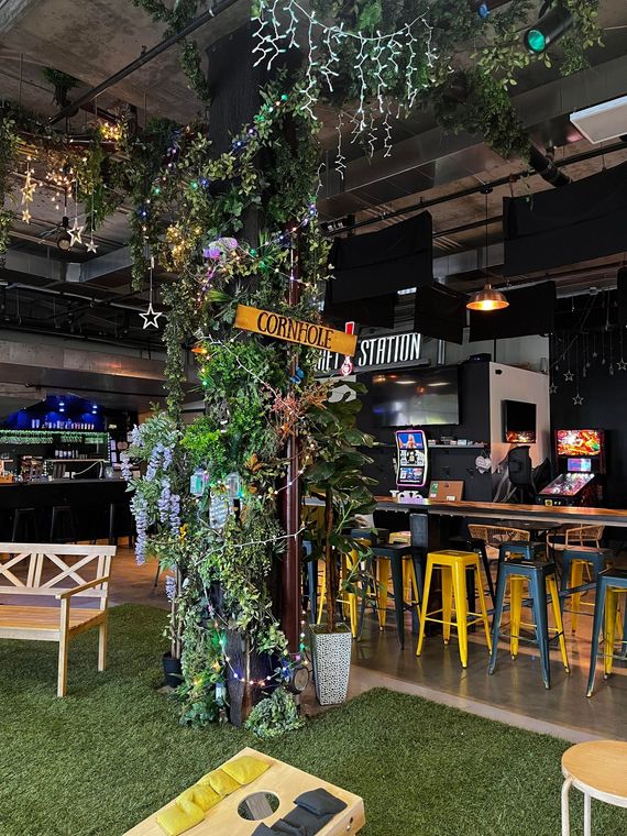 Indoor bar with artificial grass, a decorated tree, and seating. Yellow stools and arcade games are visible.