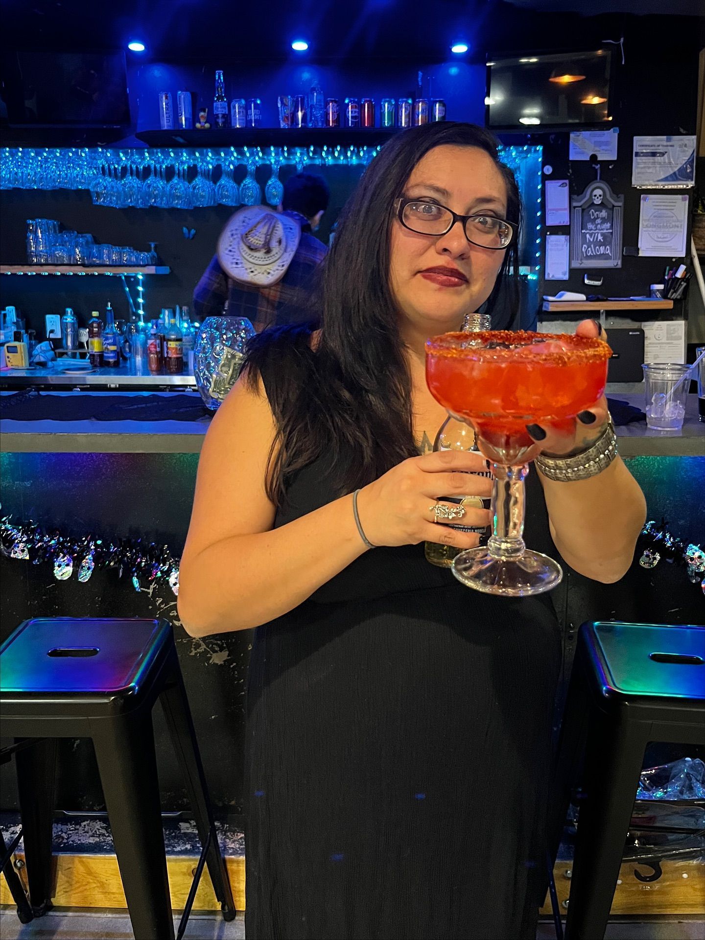 Woman holding a large red cocktail at a bar, wearing glasses and a black dress. Blue neon lights in background.