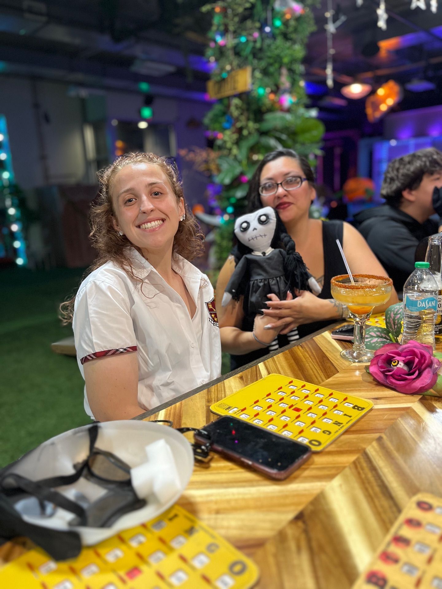 Two women at a bar; one holds a doll, a cocktail, and smiles. Colorful decor and drinks surround them.