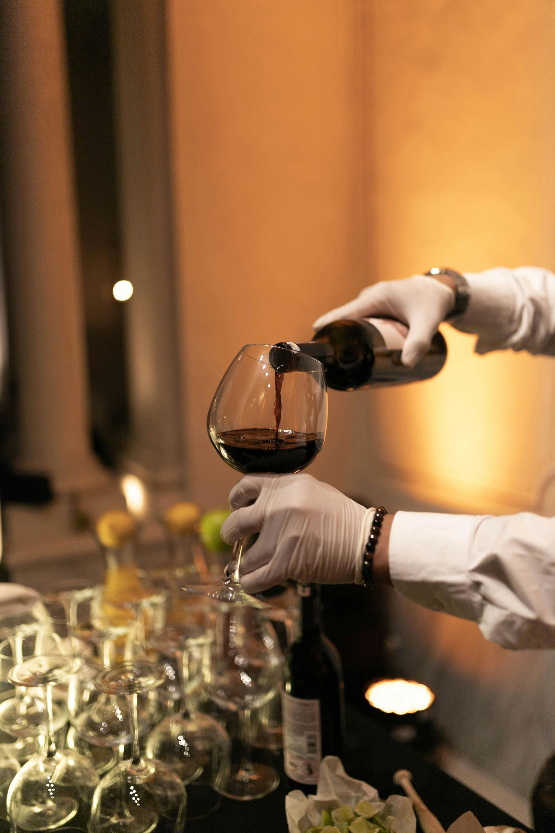 A person in gloves pours red wine into a glass, other glasses and a bottle are in the foreground, dim setting.