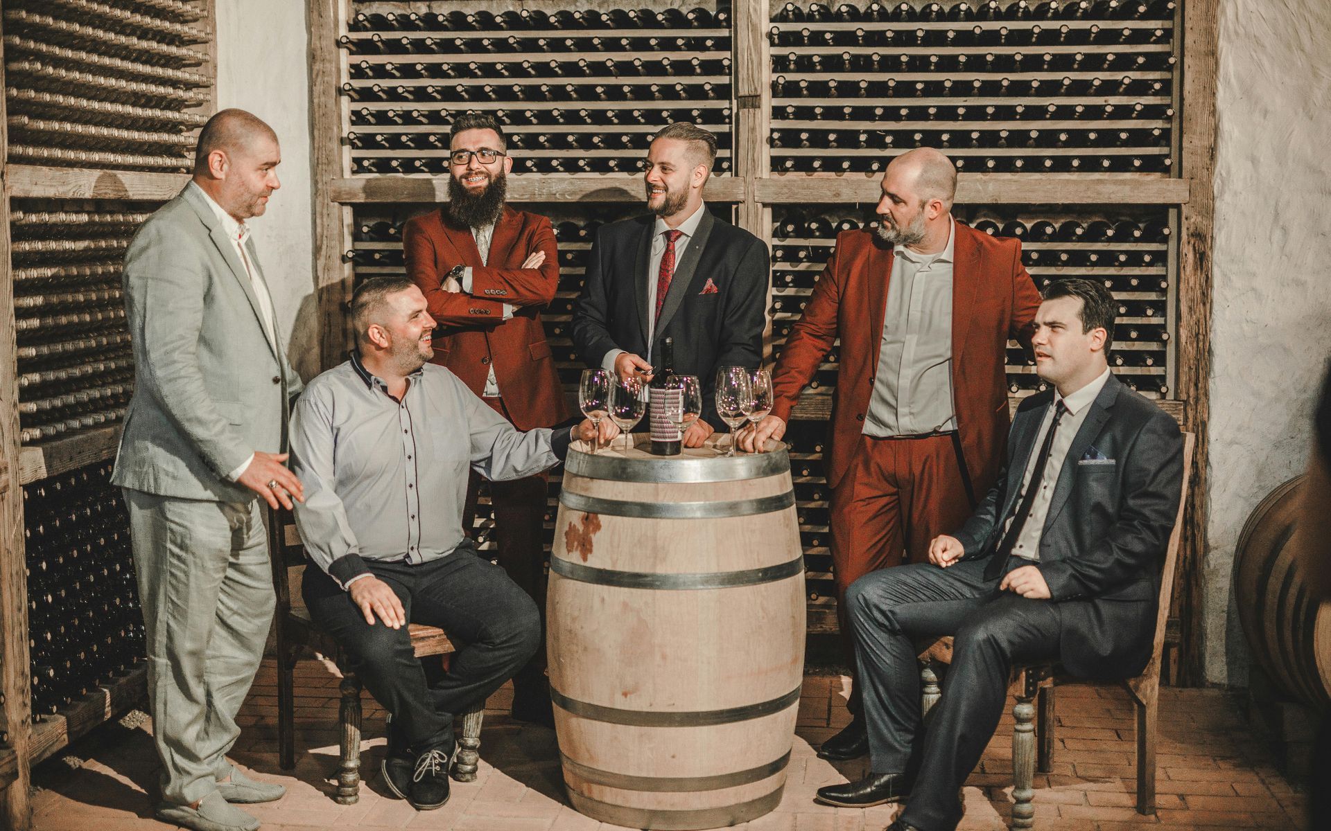 Six men in a wine cellar, some in suits, around a barrel with wine glasses.