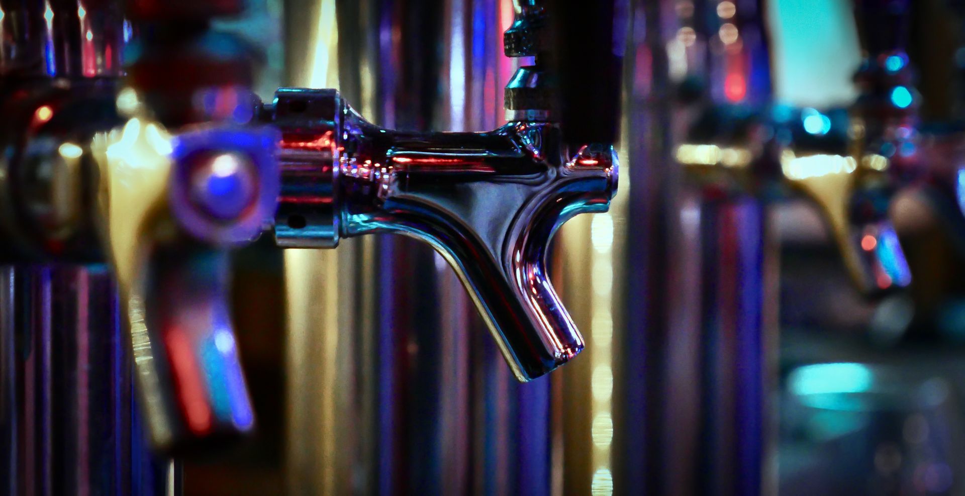Close-up of shiny silver beer taps in a dimly lit bar, with colorful neon lighting.