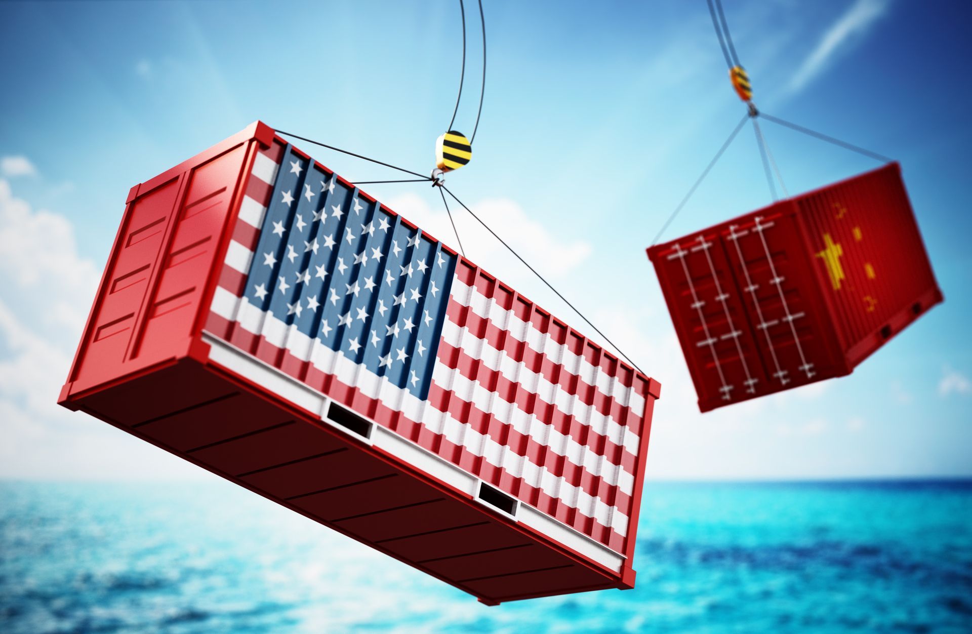 An american and a chinese shipping container are being lifted by a crane over the ocean.