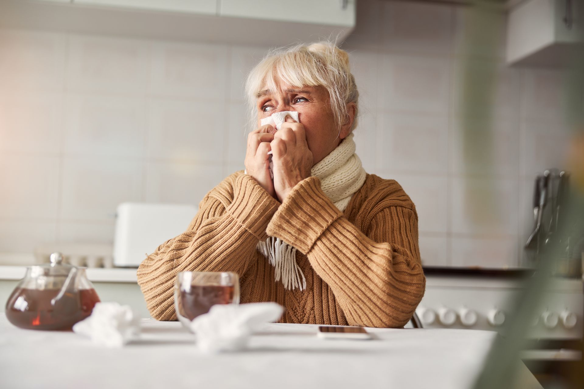 An elderly woman is blowing her nose into a napkin while sitting at a table.