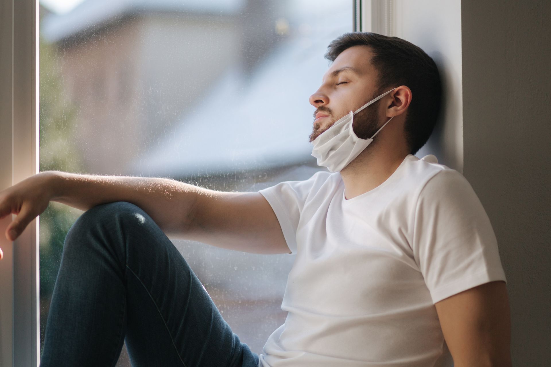 A man wearing a face mask is sitting on the floor looking out of a window.