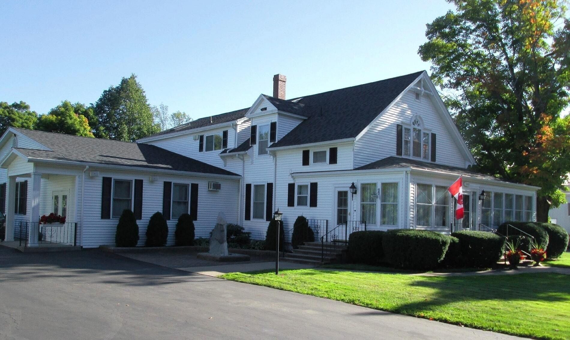 A two-story white house with dark shutters and a large wrap-around porch, surrounded by green lawns and trees.