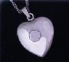 A silver heart-shaped pendant on a chain, featuring a small, circular recessed detail in the center.