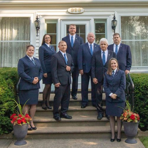 A group of eight professionals in matching blue suits stand on the front steps of a building with white trim.