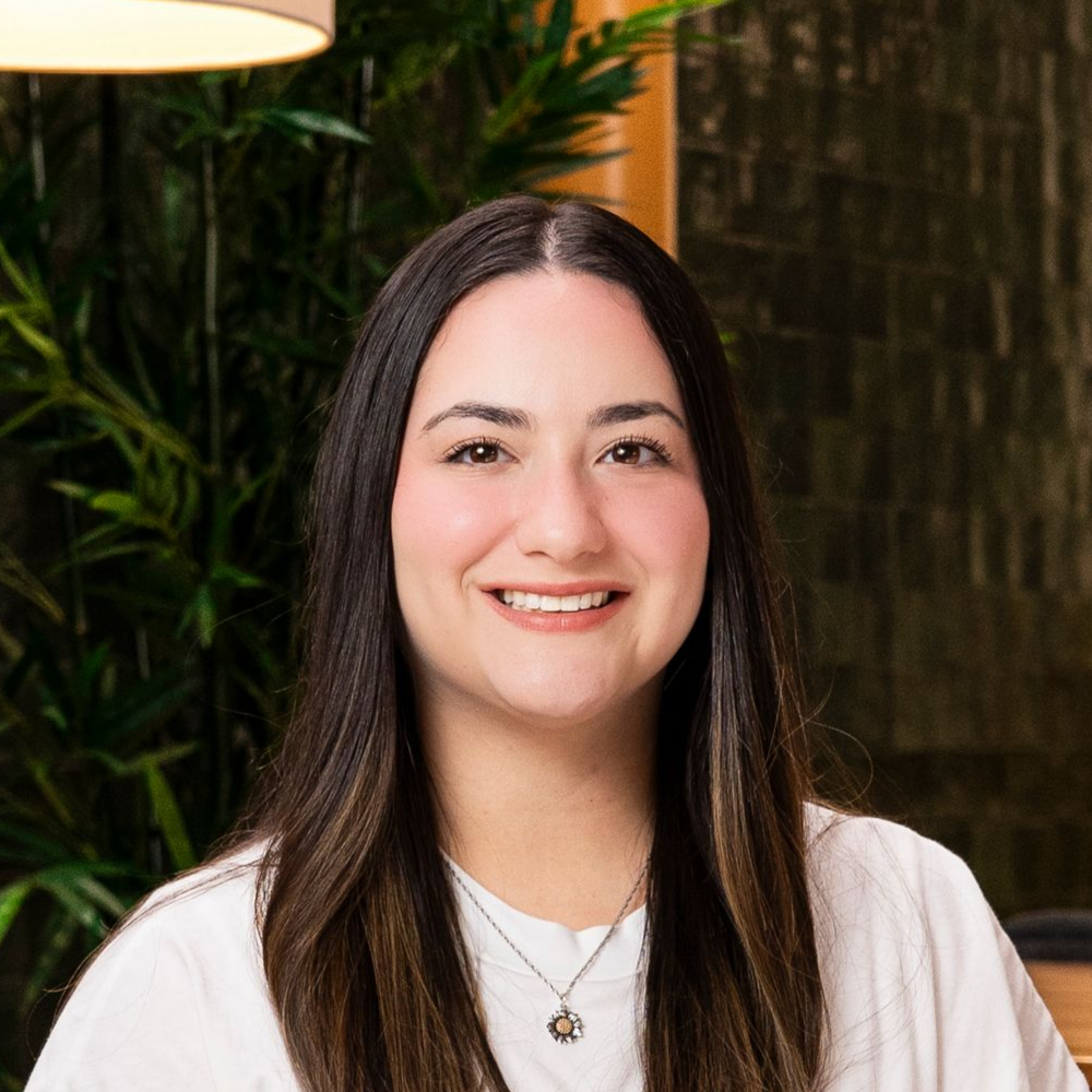 Woman with dark hair smiling, wearing a white shirt and necklace, set against a blurred background.