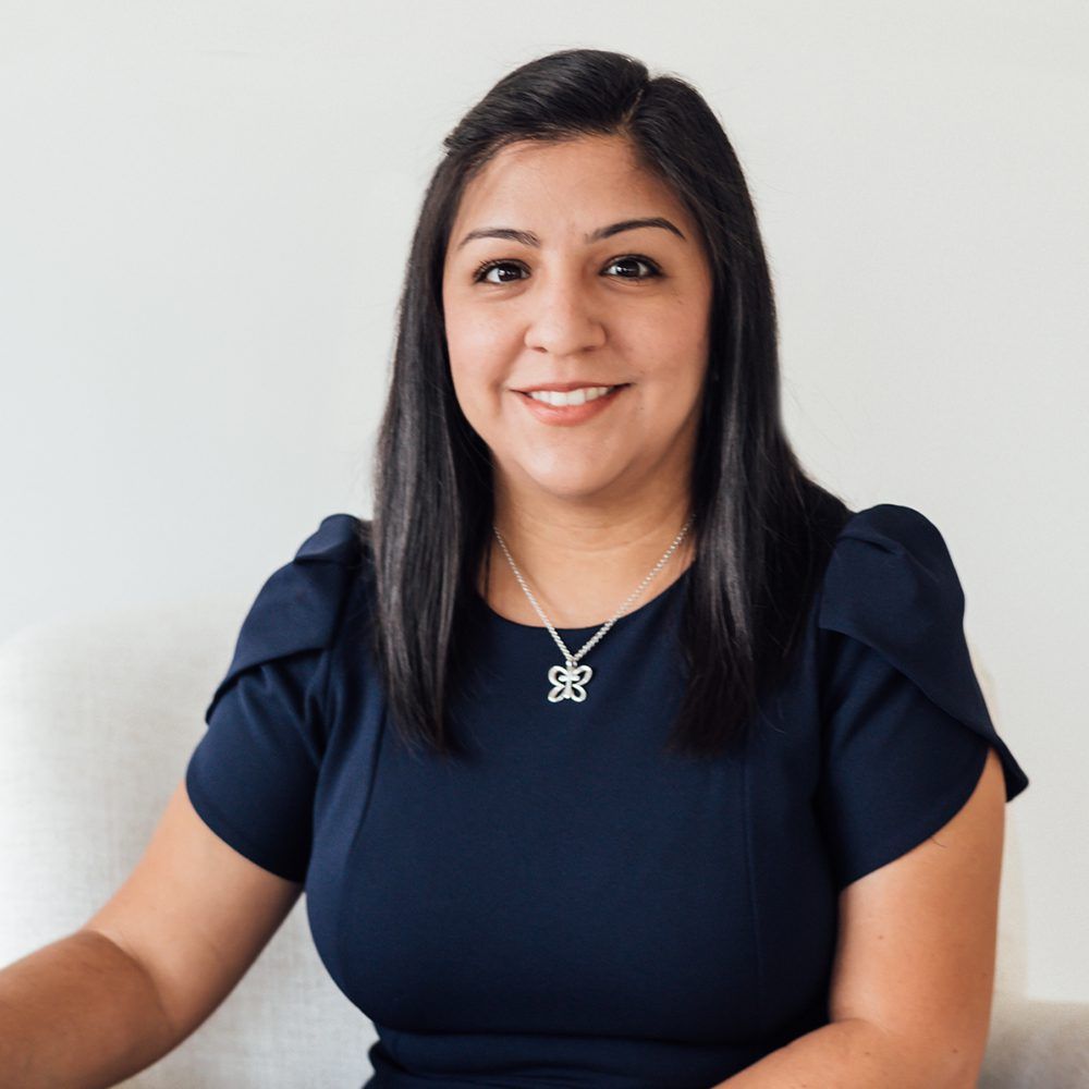 Woman with dark hair smiles, wearing a navy dress and silver necklace, seated against a white background.