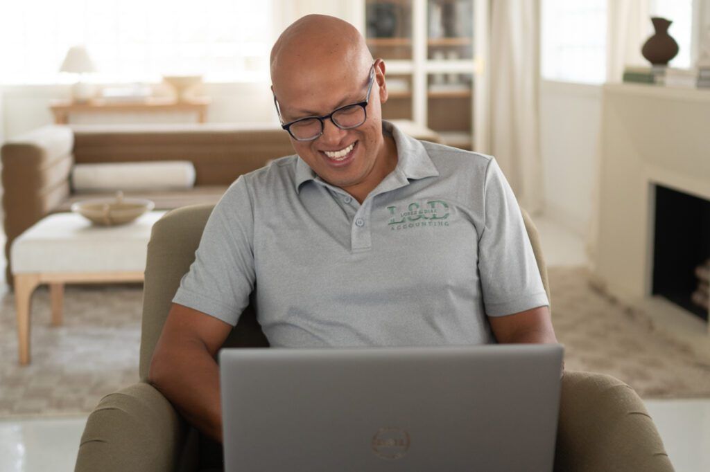 Man smiling while looking at a laptop, sitting in a chair in a living room.