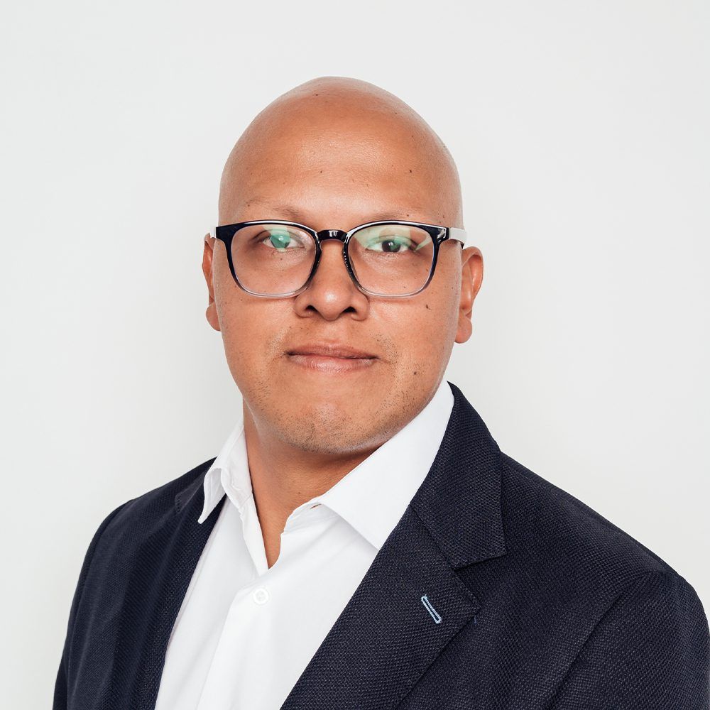 Bald man wearing glasses, white shirt, and dark blazer, looking forward against a white backdrop.