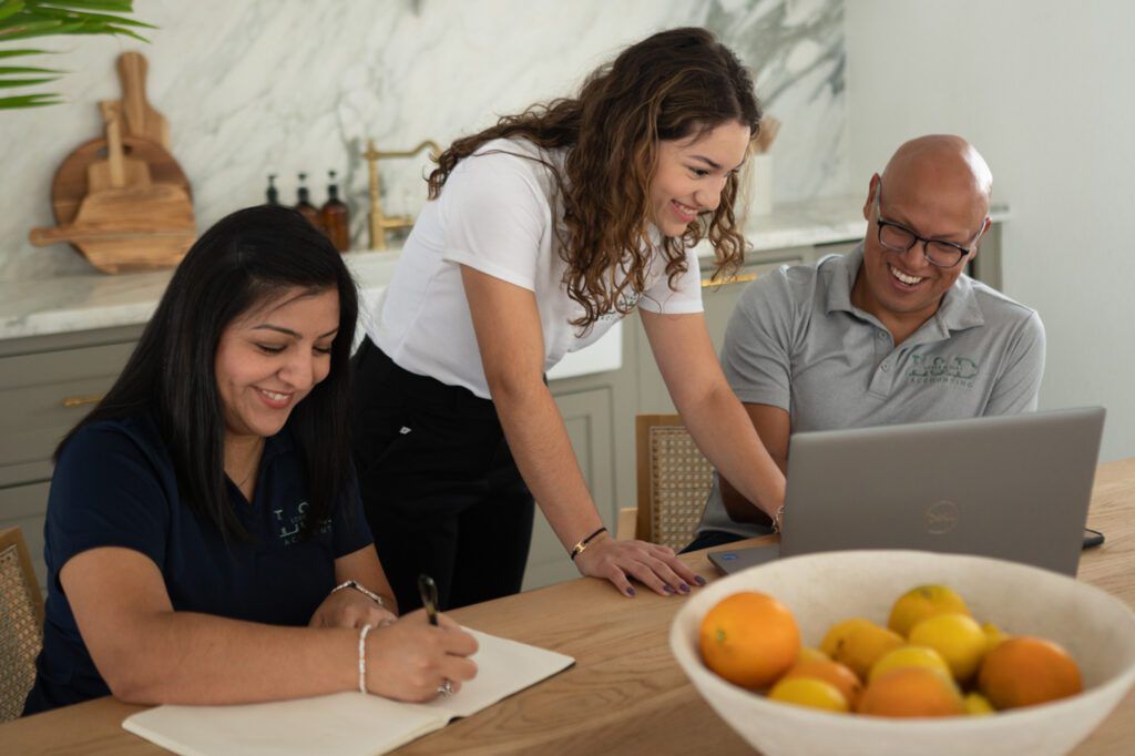 Three people collaborating around a table, one writing, one on a laptop, and the third leaning in, smiling.