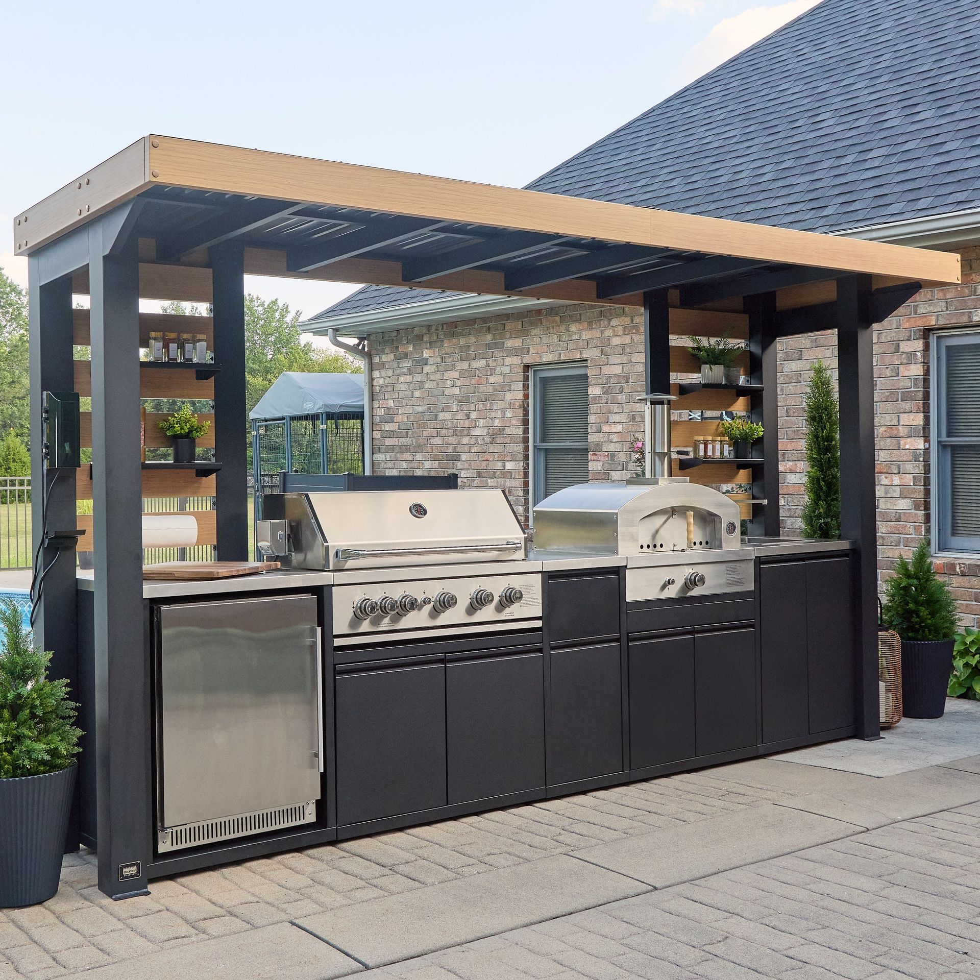 Outdoor kitchen with stainless steel appliances under a covered structure. Dark cabinets and shelves, brick backdrop.