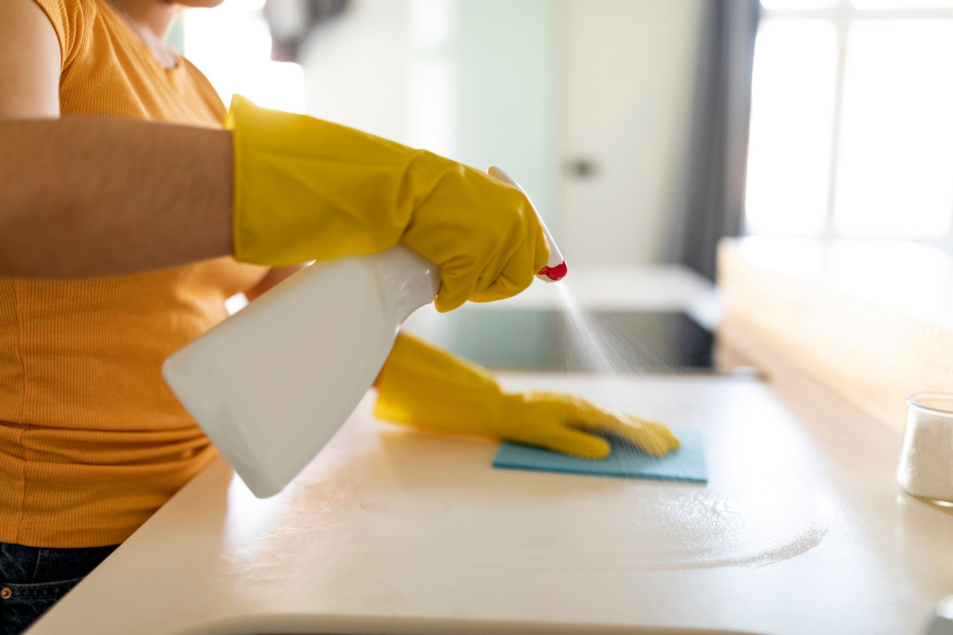 A woman wearing yellow gloves is cleaning a counter with a spray bottle.