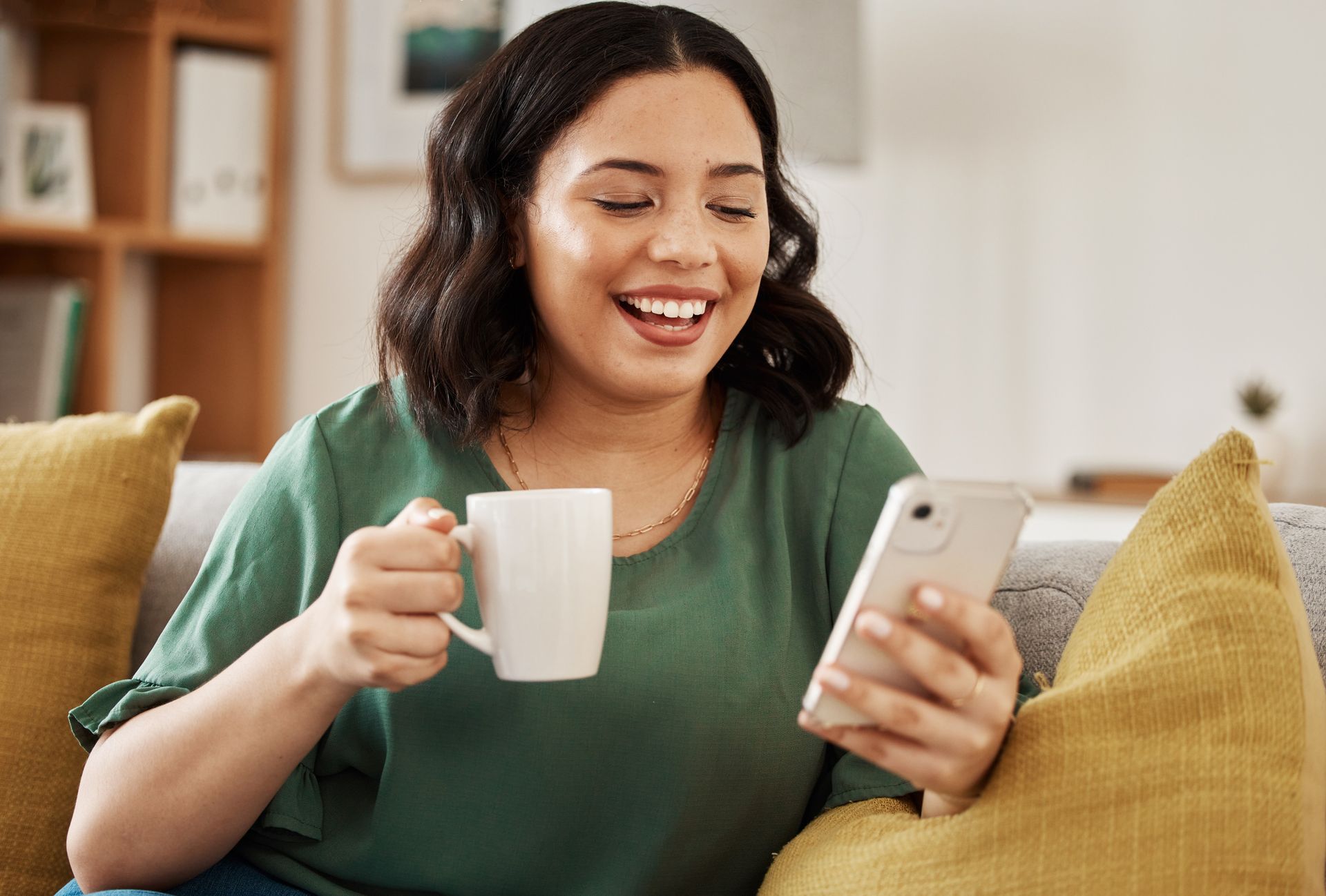 A woman is sitting on a couch holding a cup of coffee and looking at her phone.