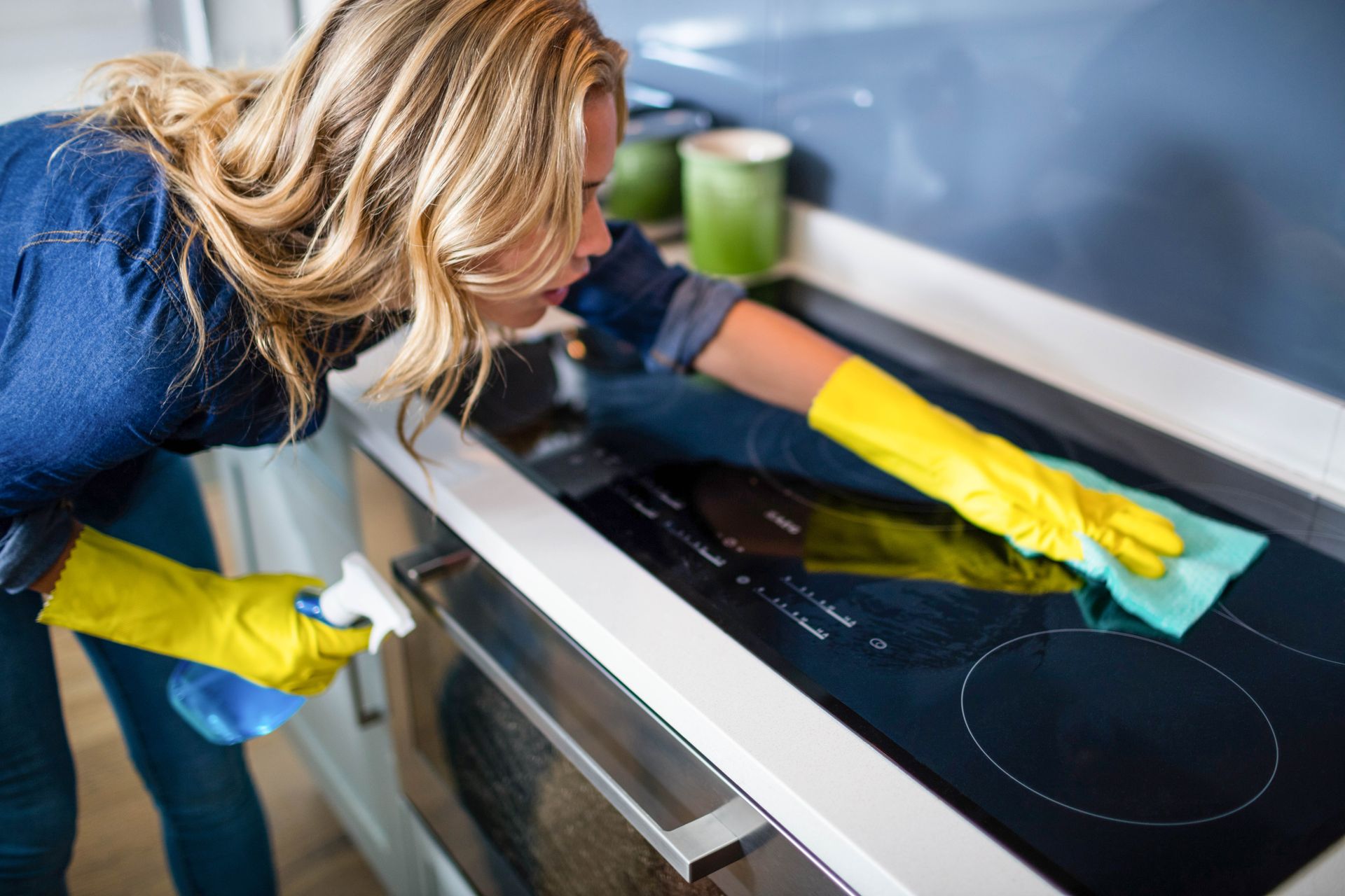 Woman in yellow gloves cleans a black stovetop with a spray bottle and blue cloth in a kitchen.