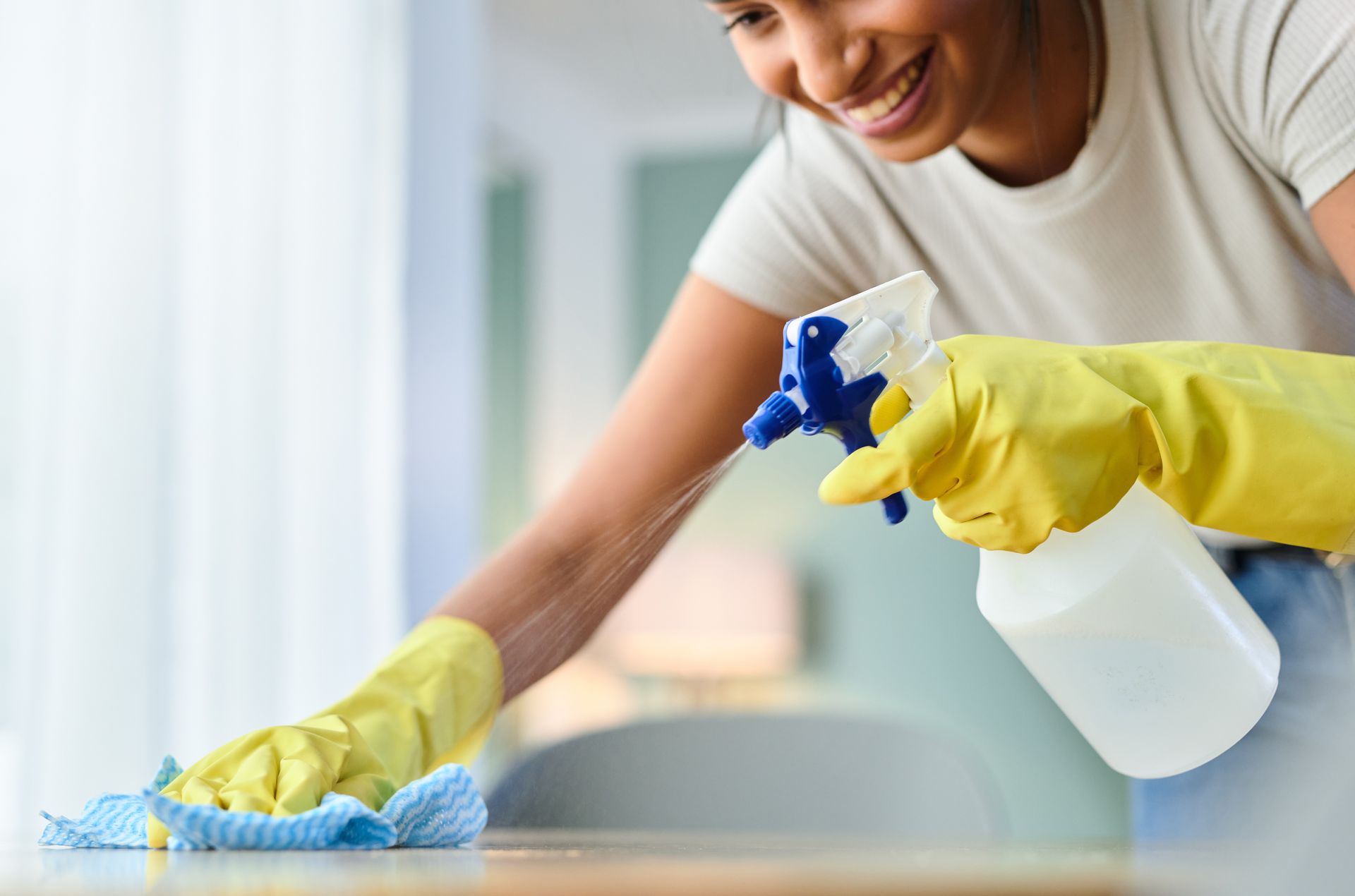Woman wearing yellow gloves cleaning a table with spray bottle and cloth, smiling.