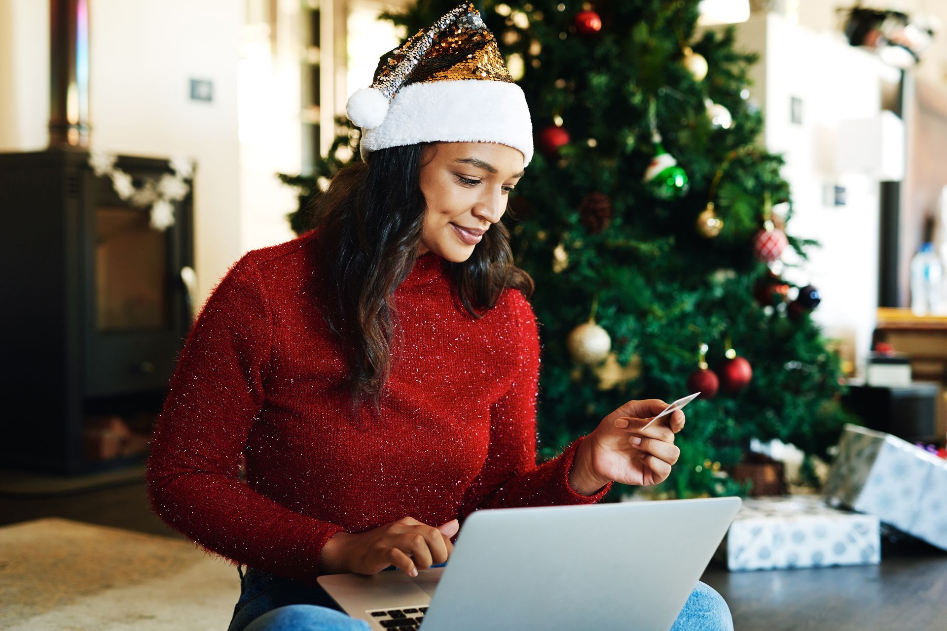 Woman in Santa hat, red sweater shopping online with laptop in front of Christmas tree, holding credit card.