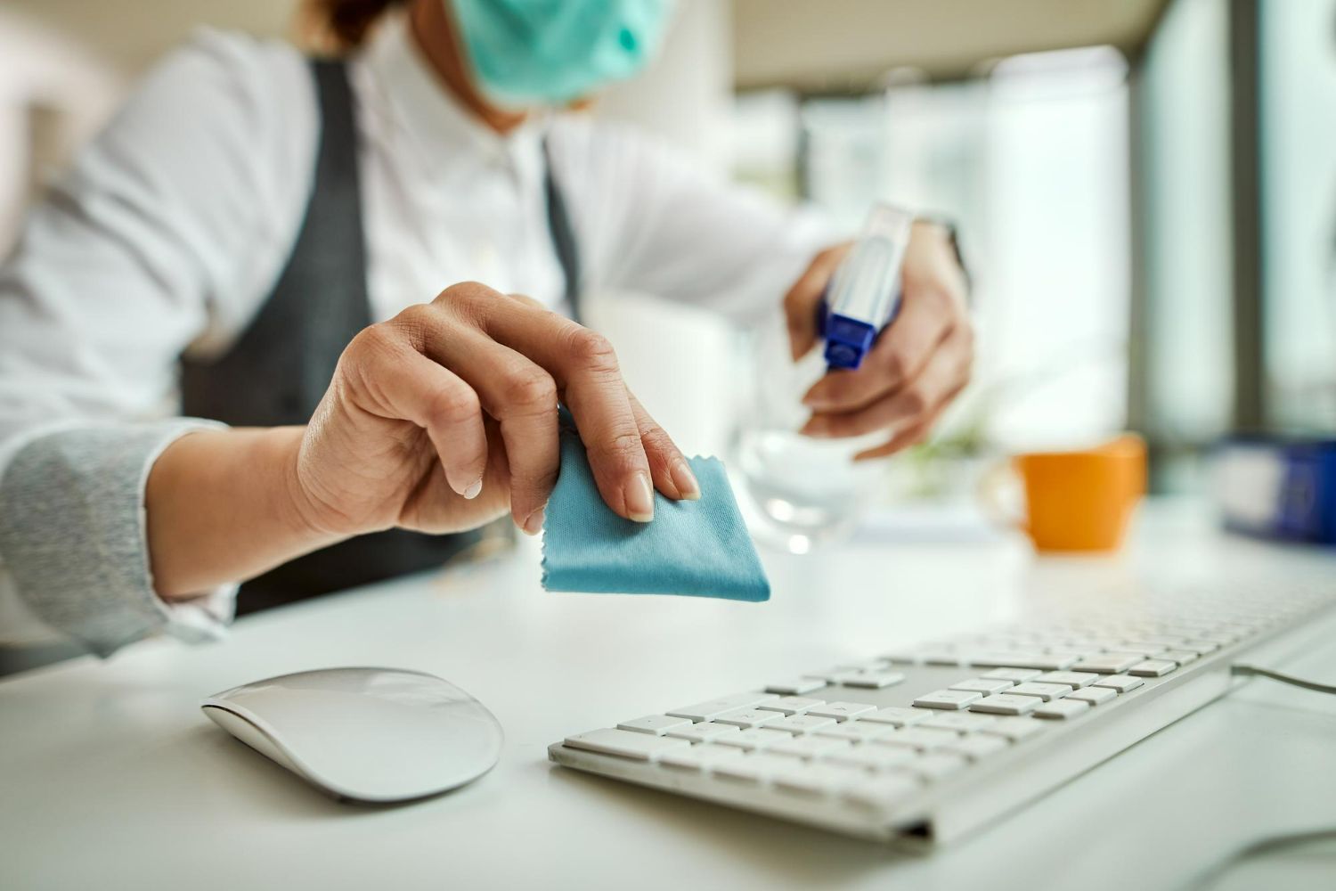 A woman wearing a mask is cleaning a computer keyboard with a cloth.