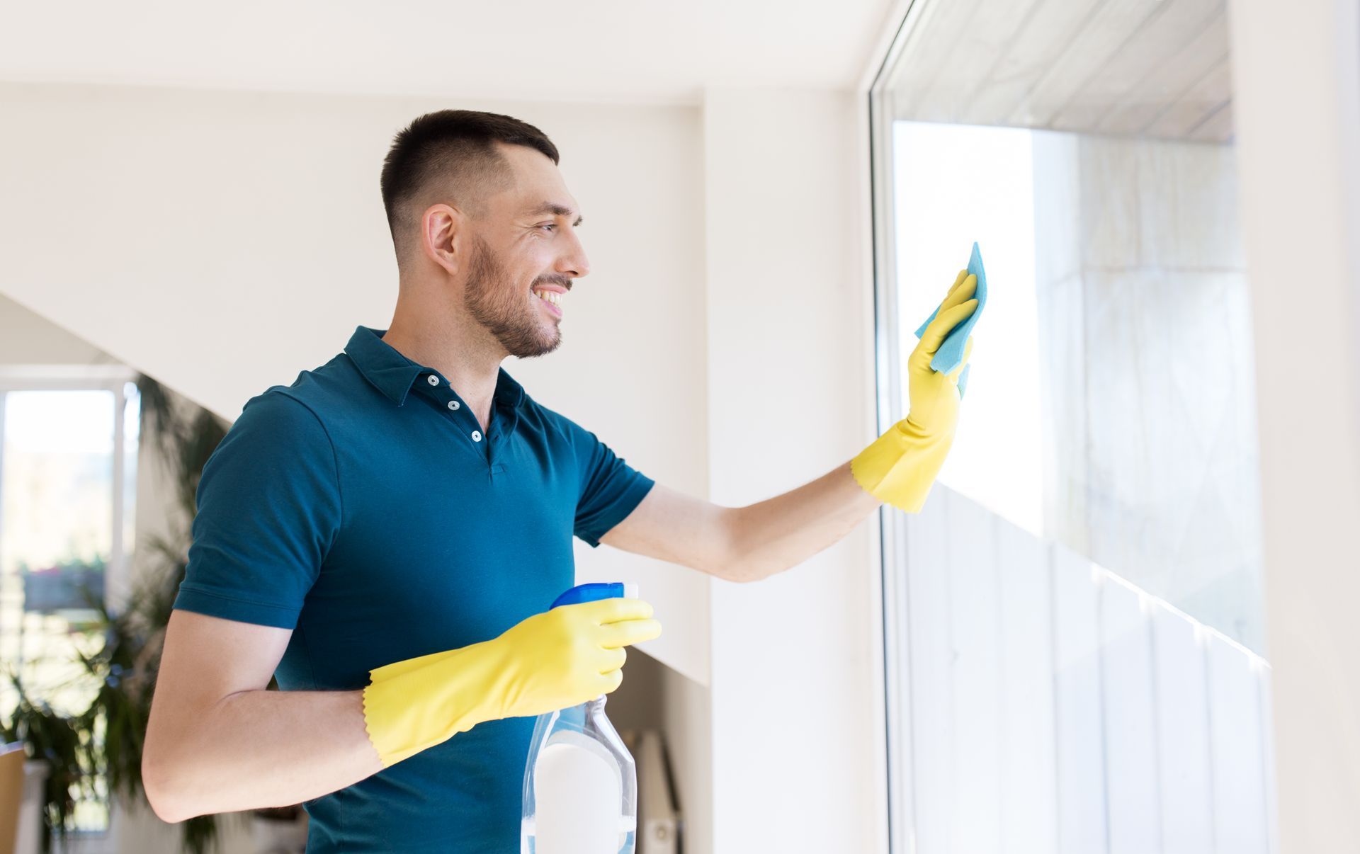 Man wearing yellow gloves cleaning a window with a spray bottle; smiling in a bright room.