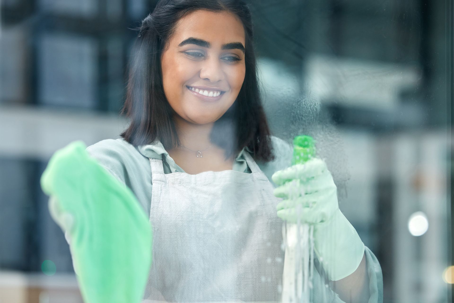 Woman wearing gloves and apron smiles while spraying and wiping a window.