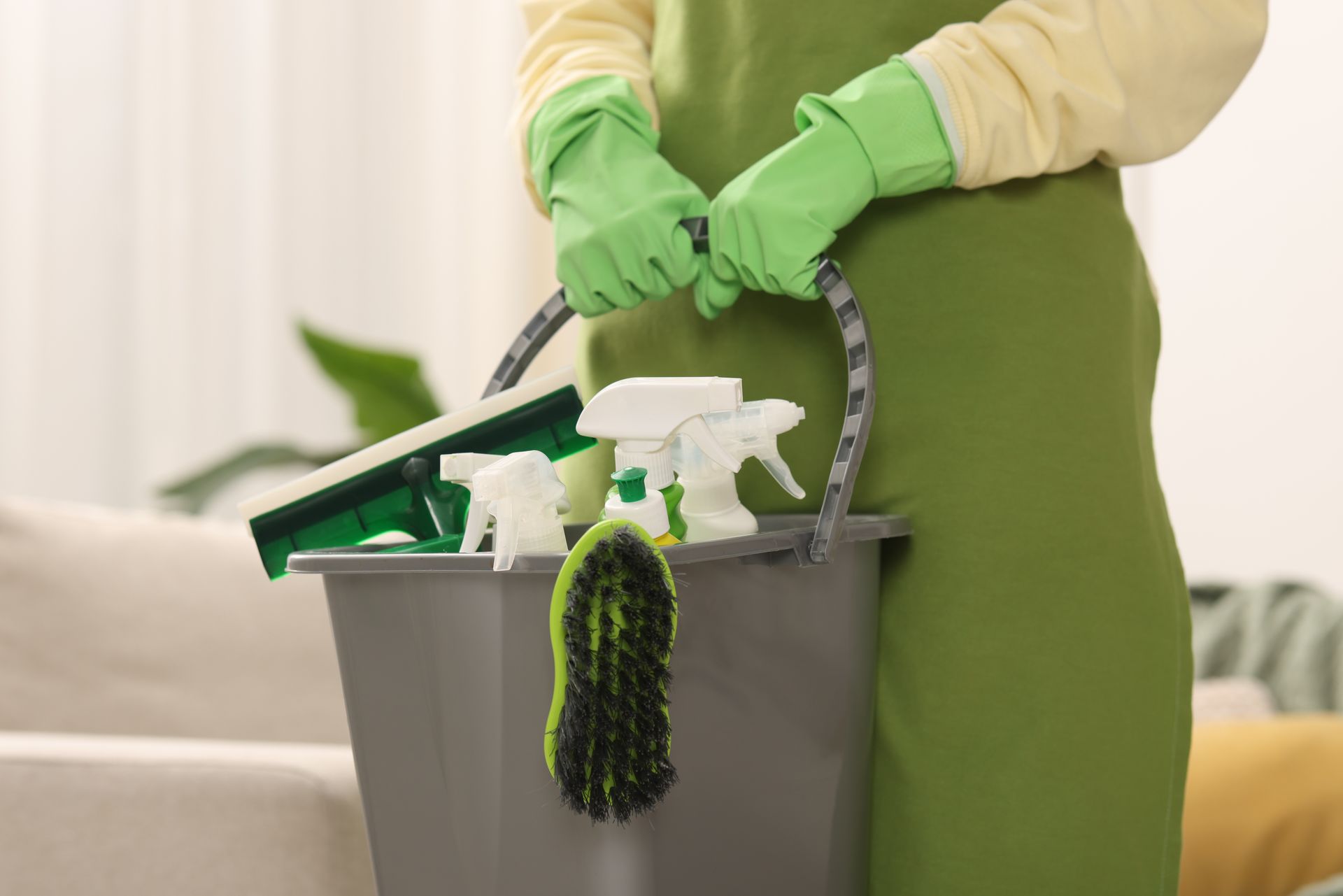 Person in green apron and gloves holding a bucket with cleaning supplies: spray bottles, brush, and squeegee.