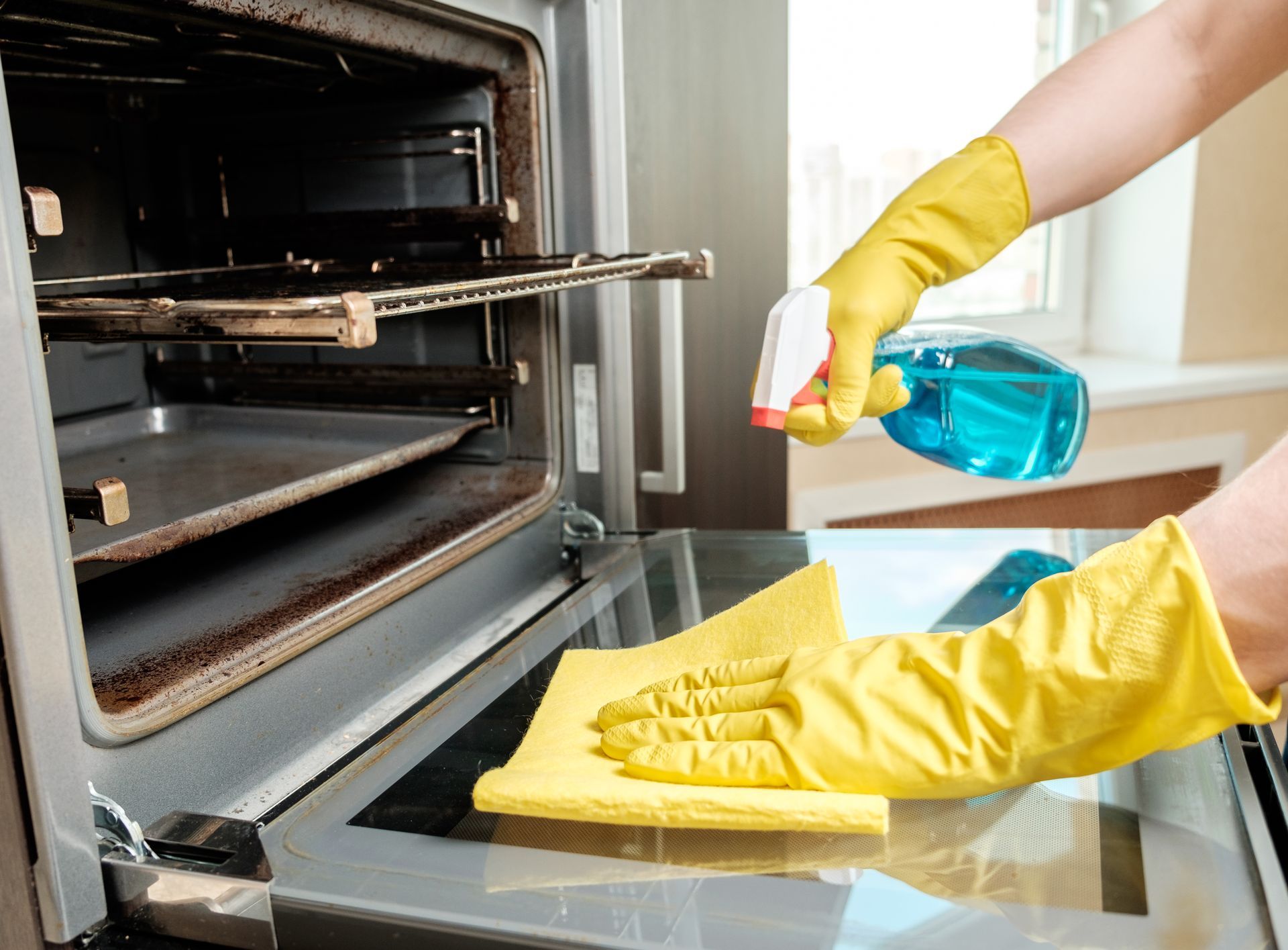 A person wearing yellow gloves is cleaning an oven with a cloth and spray bottle.