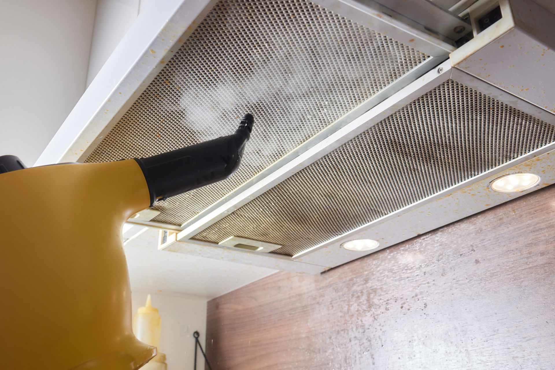 A person uses a steam cleaner on a dirty range hood filter in a kitchen.