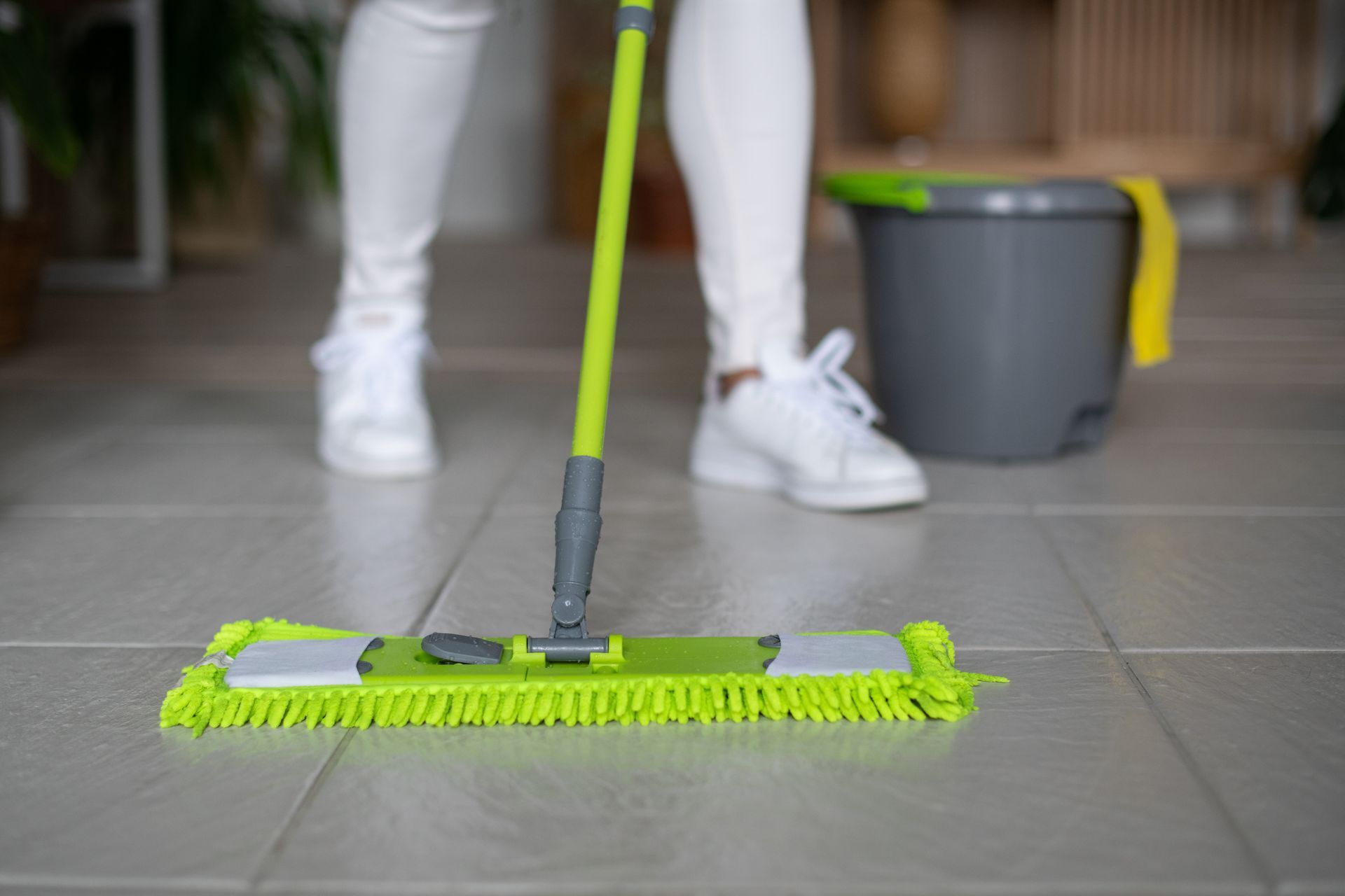 Person mopping a tiled floor with a green mop, a bucket is beside them.