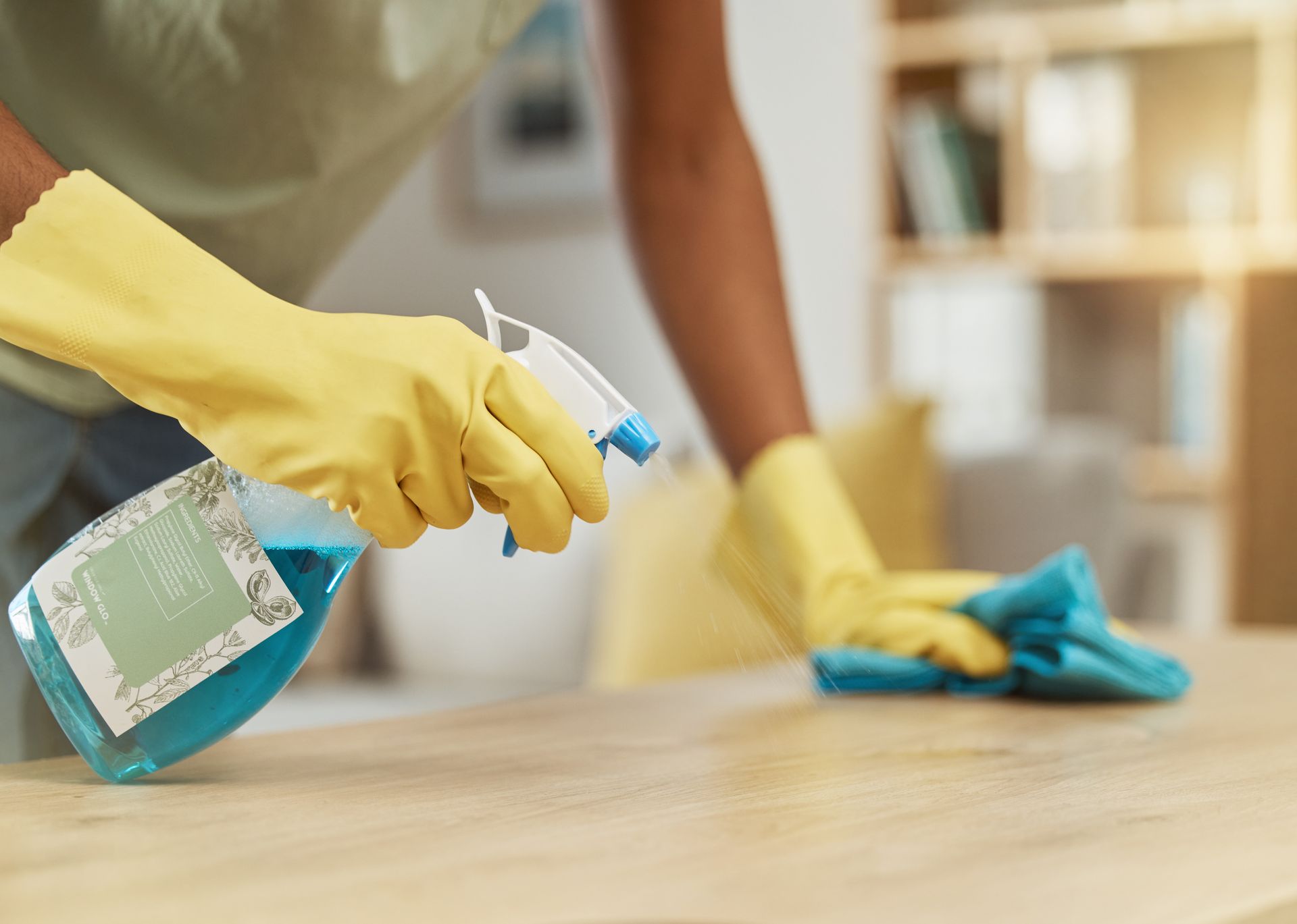 A person wearing yellow gloves is cleaning a table with a spray bottle and a cloth.