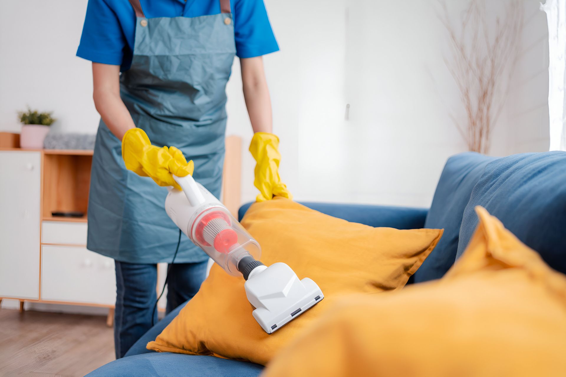 Person vacuuming a blue sofa with a yellow cover, wearing gloves and an apron in a bright living room