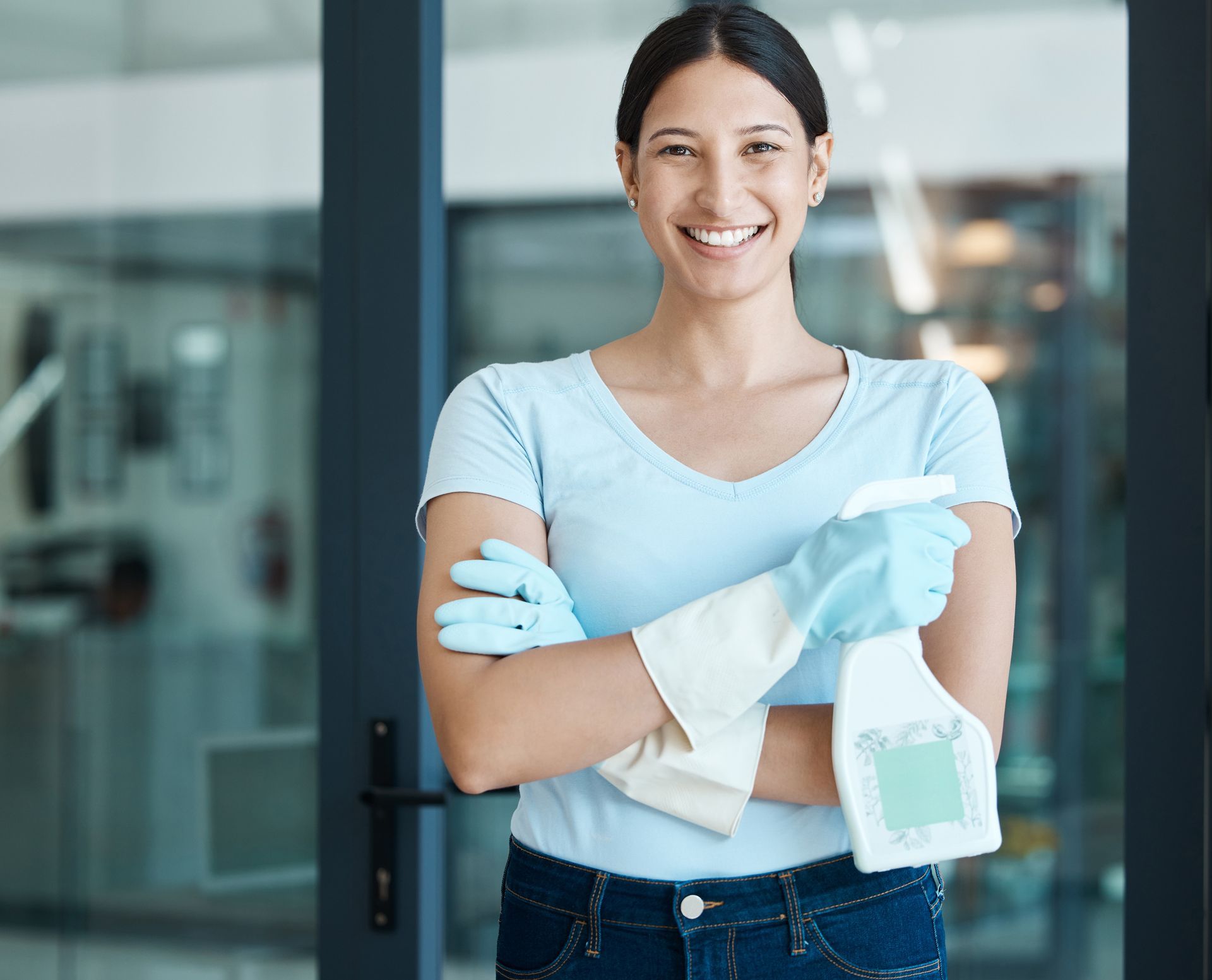 Woman in blue gloves and shirt holds cleaning spray, smiling.