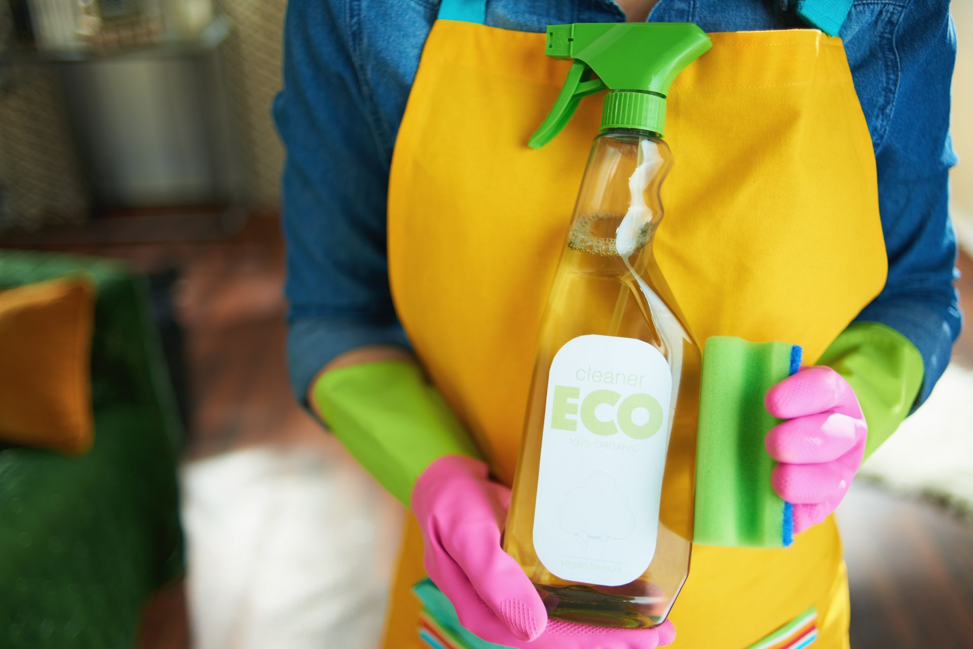 A woman in a yellow apron and pink gloves is holding a bottle of eco cleaner.