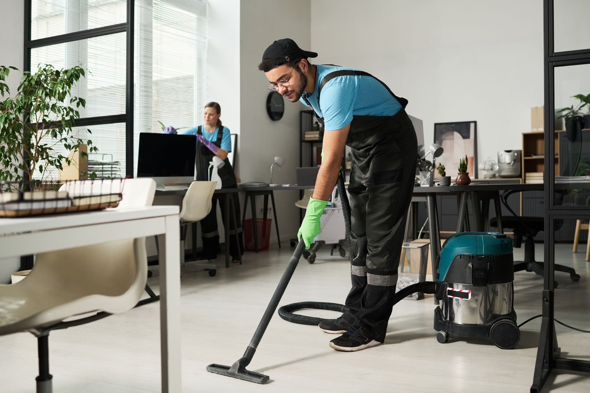 Man vacuuming office floor, wearing overalls, other person dusting in background.