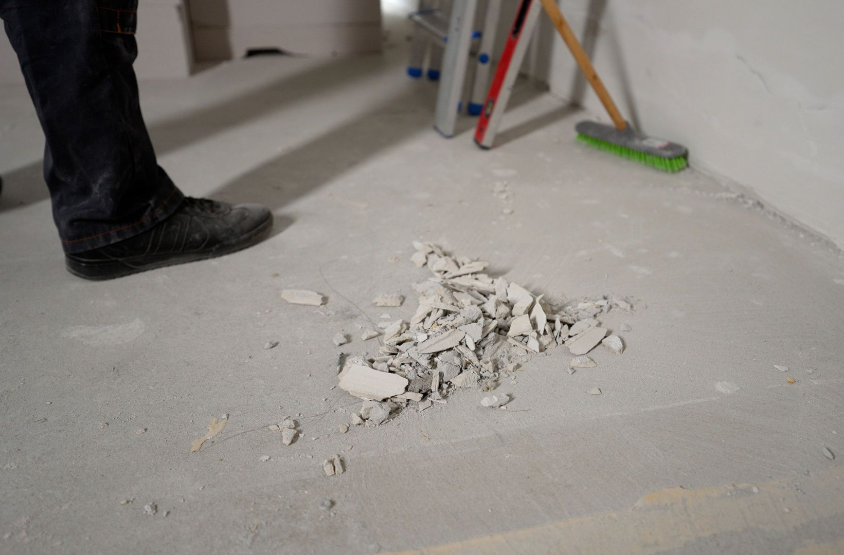 Small pile of white plaster or debris on a dusty floor beside a person’s boot and a ladder