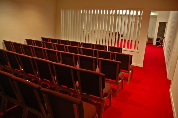 Rows of chairs in a room with red carpet, leading to a hallway. White blinds and walls.